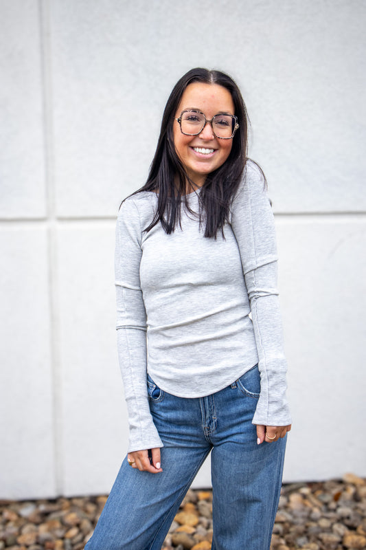 Woman wearing a gray sweater and blue jeans standing against a white wall.