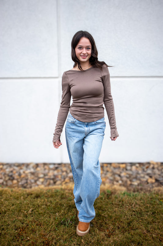 Woman wearing a brown long-sleeve top and light blue jeans standing outdoors.