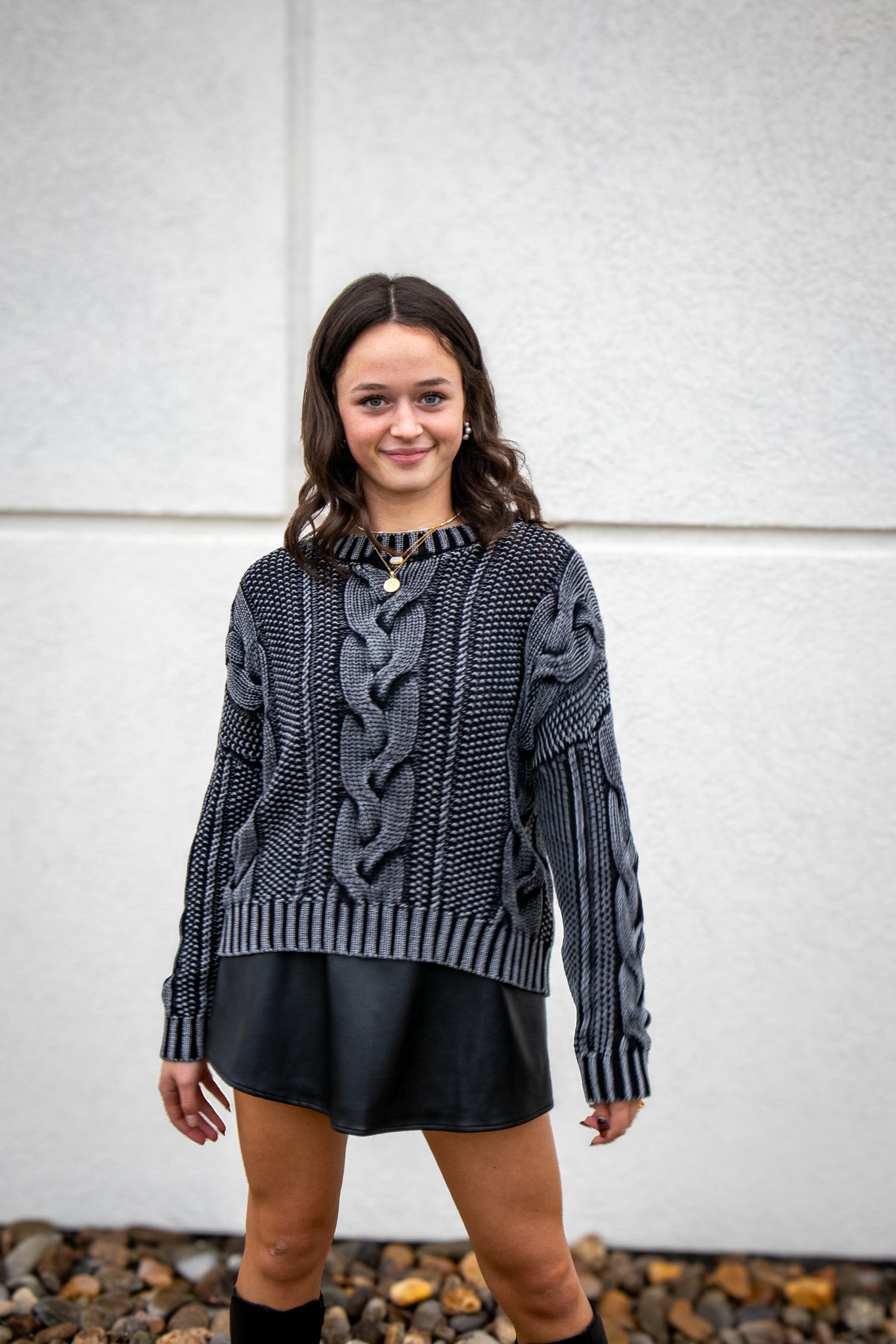 Woman wearing a cable knit sweater and black skirt against a white wall.