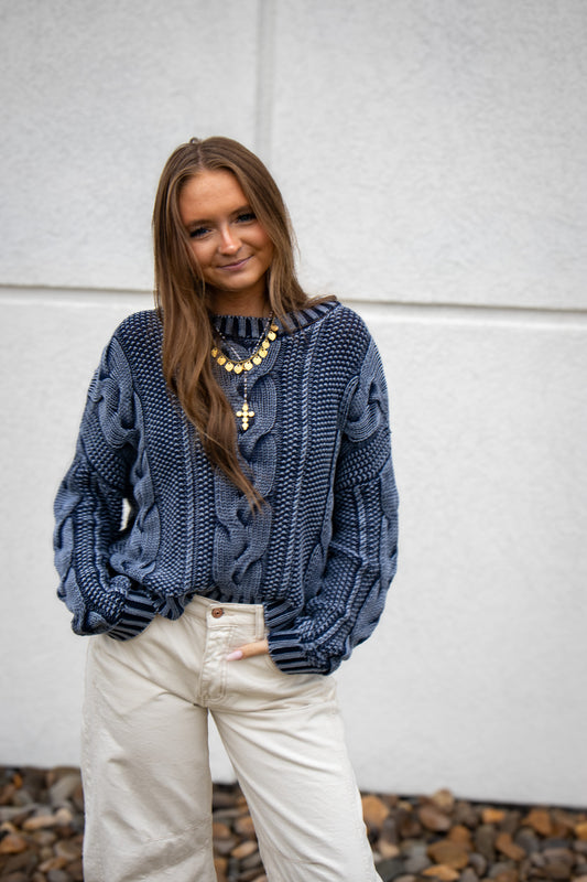 Woman wearing a blue cable knit sweater and white pants against a light gray wall.