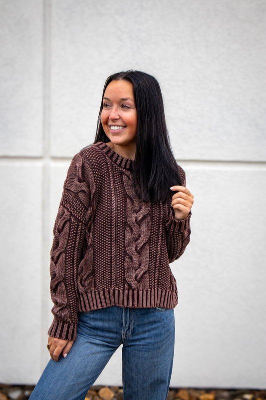 Woman wearing a brown cable knit sweater and blue jeans against a white wall.