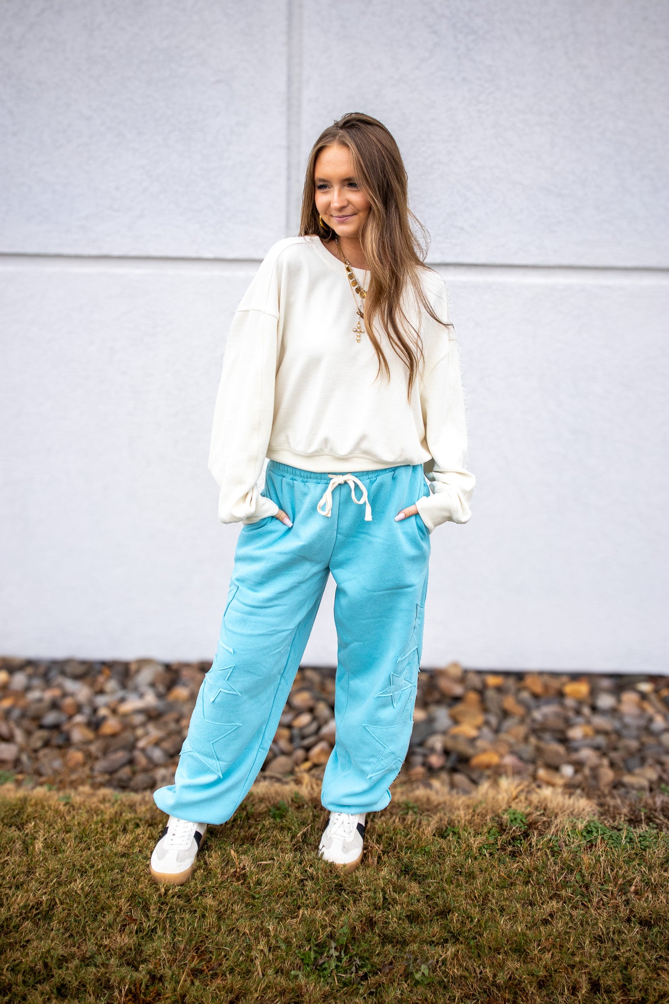 Woman wearing a white sweatshirt and blue pants standing outdoors against a gray wall.