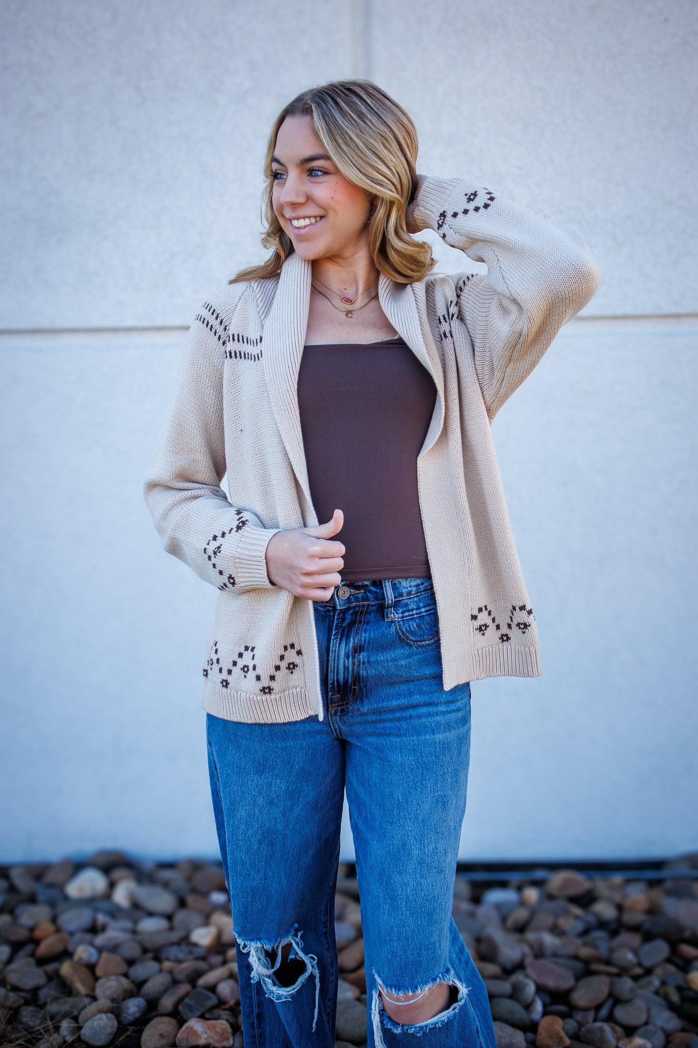 Woman wearing a beige cardigan with decorative cutouts, brown top, and blue jeans against a white wall.