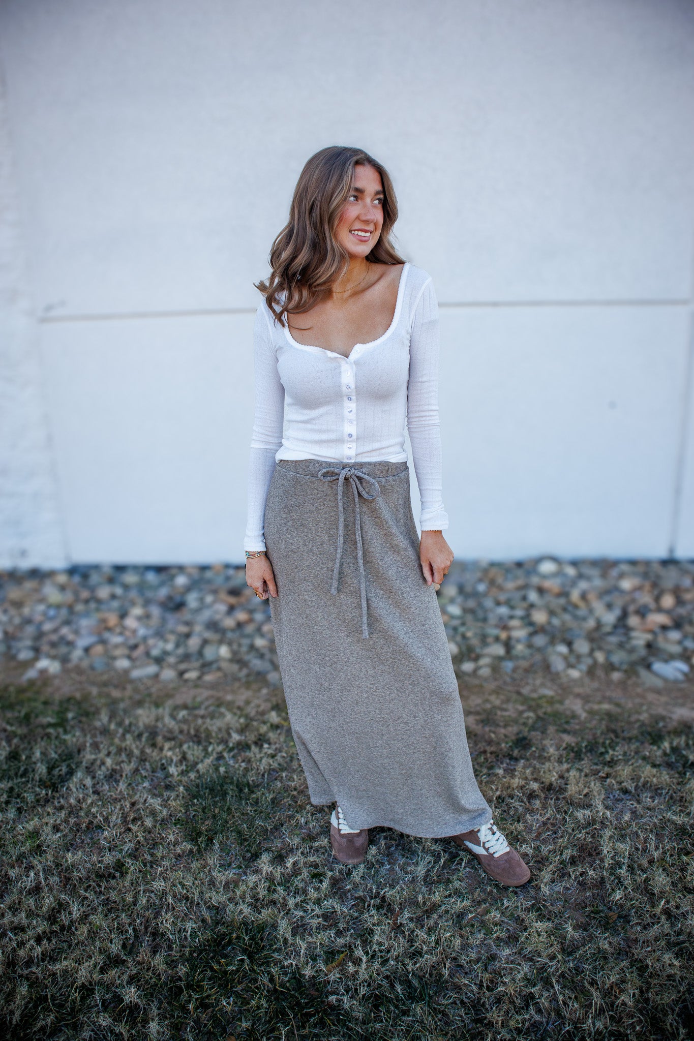 Woman wearing a white long-sleeve top and gray patterned skirt standing against a white wall.