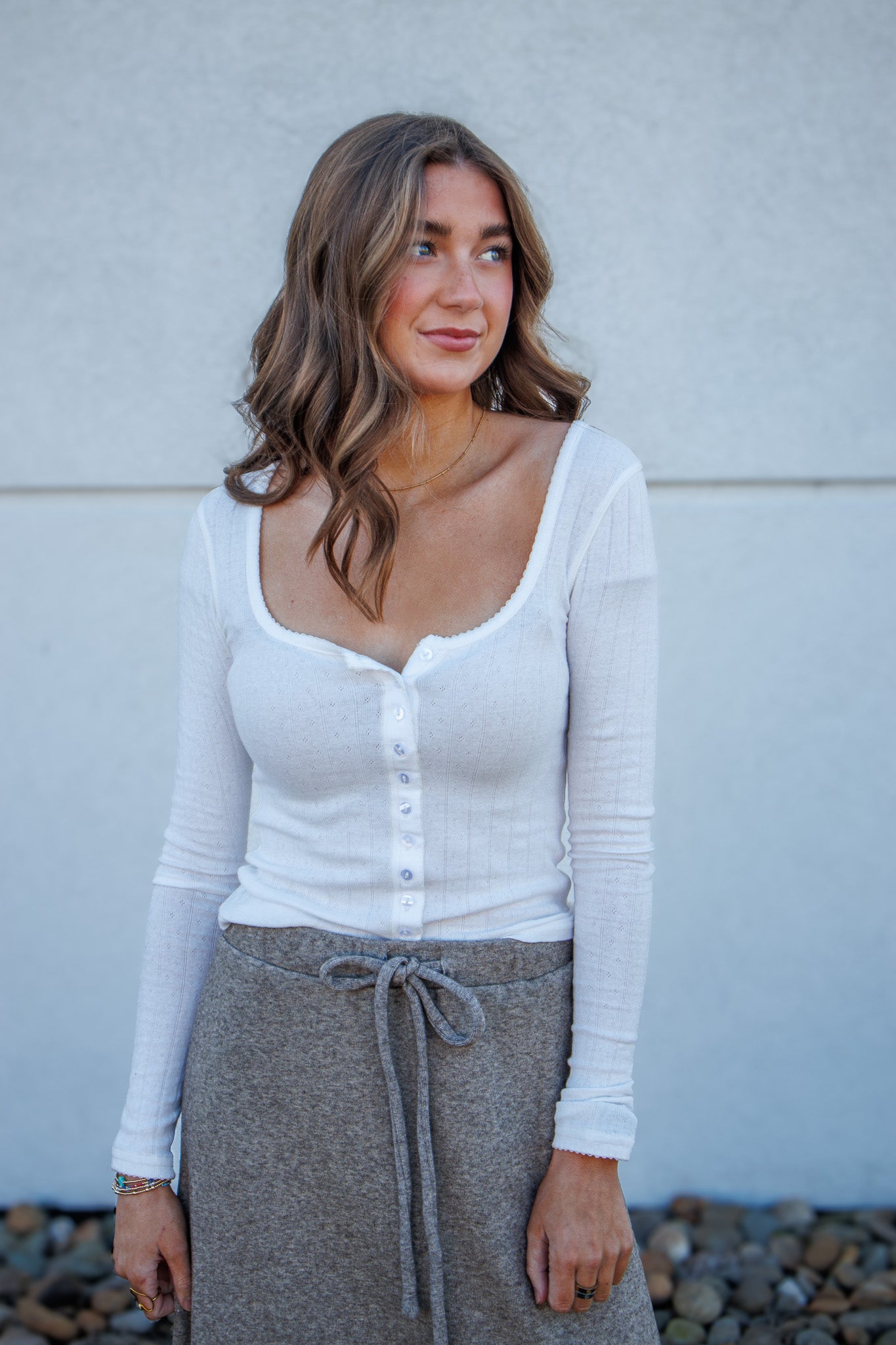 Woman wearing a white long-sleeve top and gray skirt against a light gray wall.