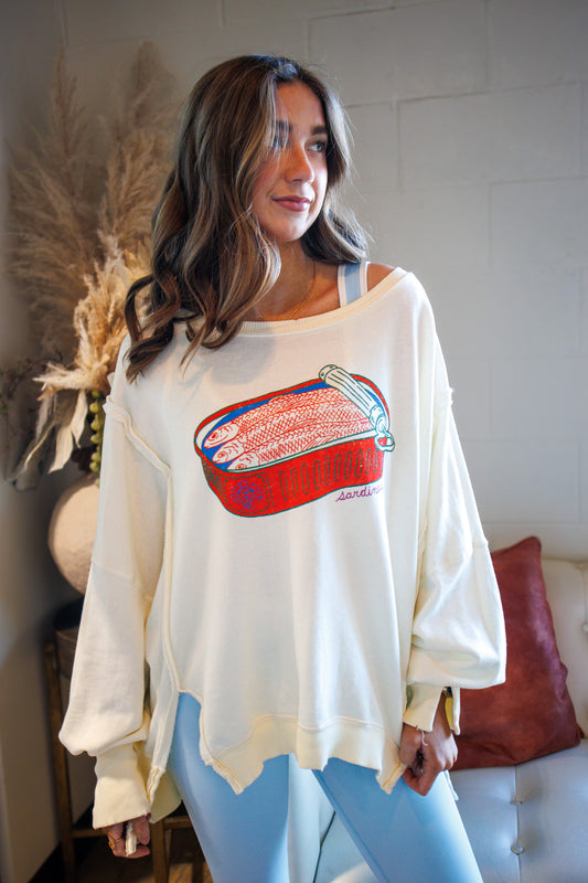 Woman wearing a white oversized sweatshirt with a graphic design, standing in a room with a white wall and decorative elements.