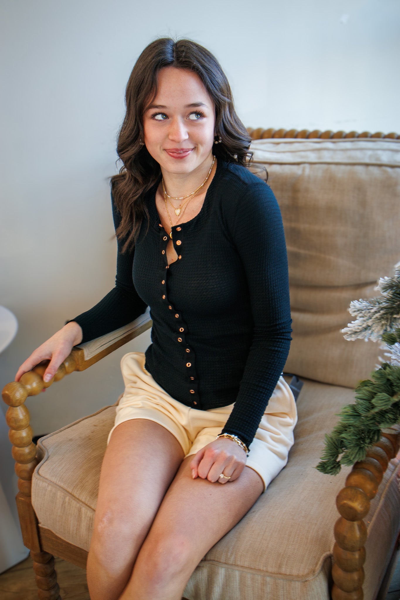 Woman sitting on a beige chair wearing a black cardigan and yellow shorts, with a neutral background.