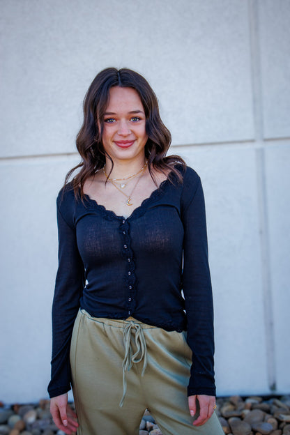 Woman wearing a black long-sleeve top with lace detail and sage green pants against a light gray wall.