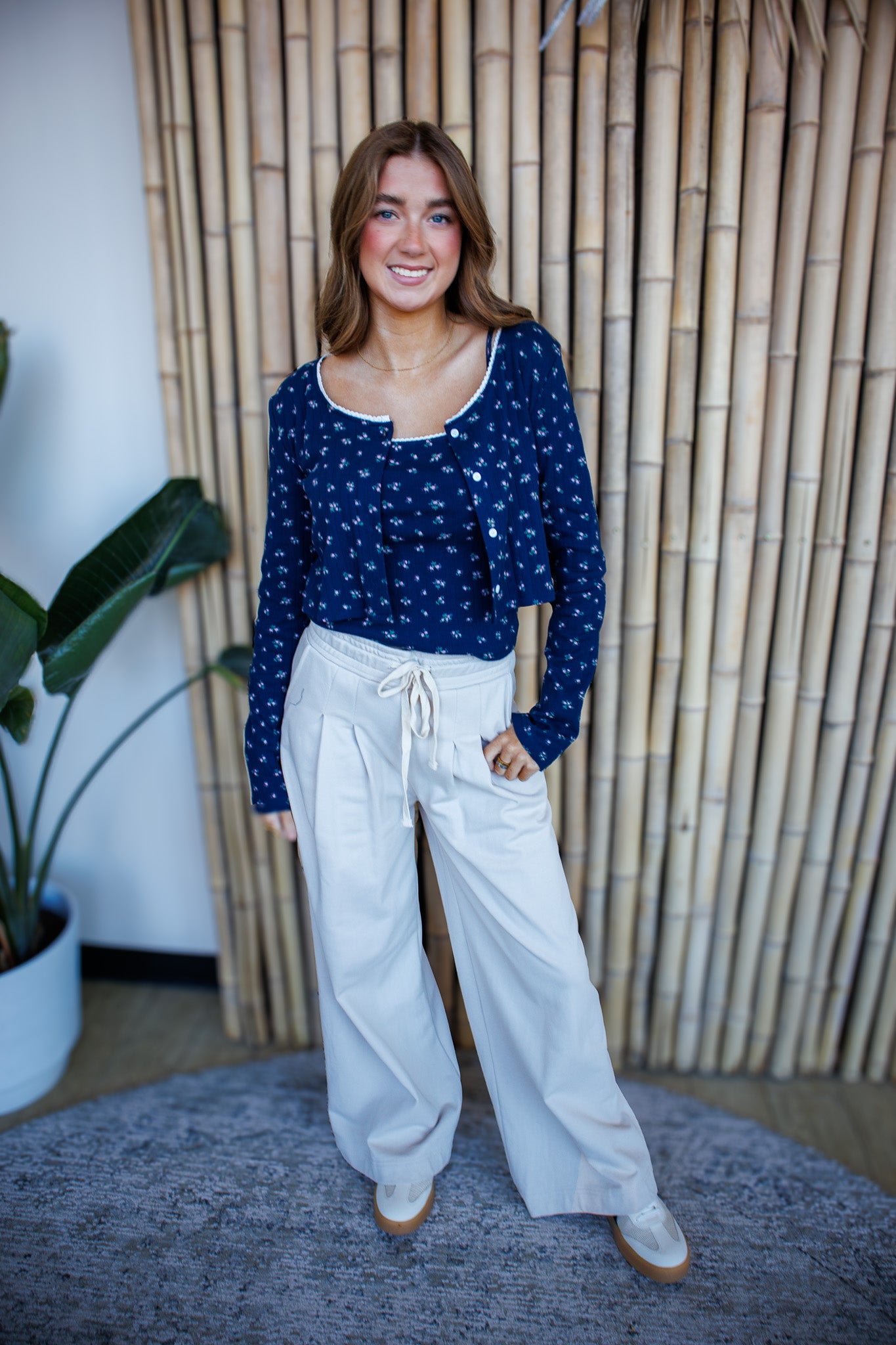 Woman wearing a blue polka dot top and white pants standing in front of a bamboo wall.