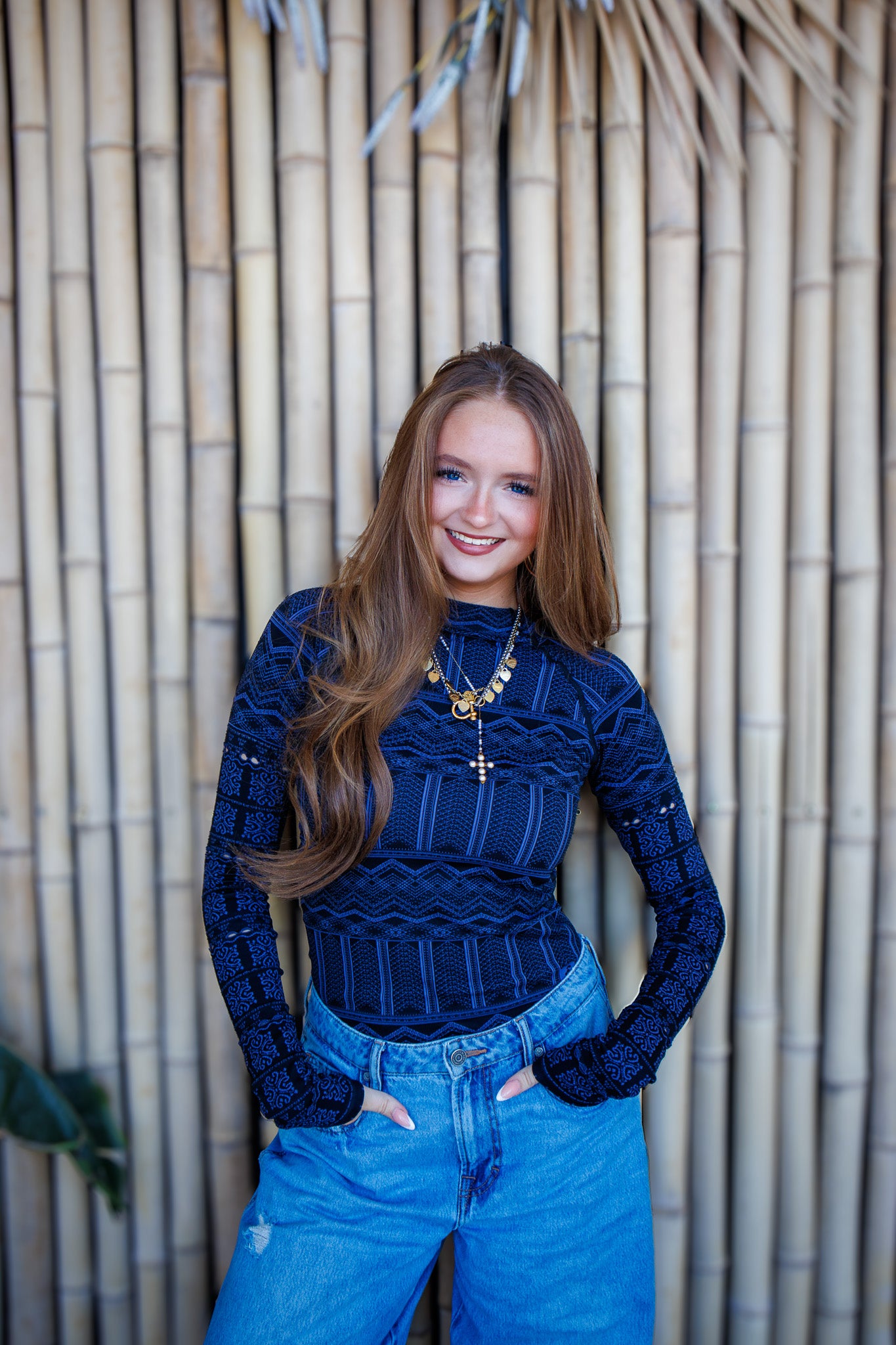 Woman wearing a blue ribbed sweater and jeans standing against a bamboo wall.