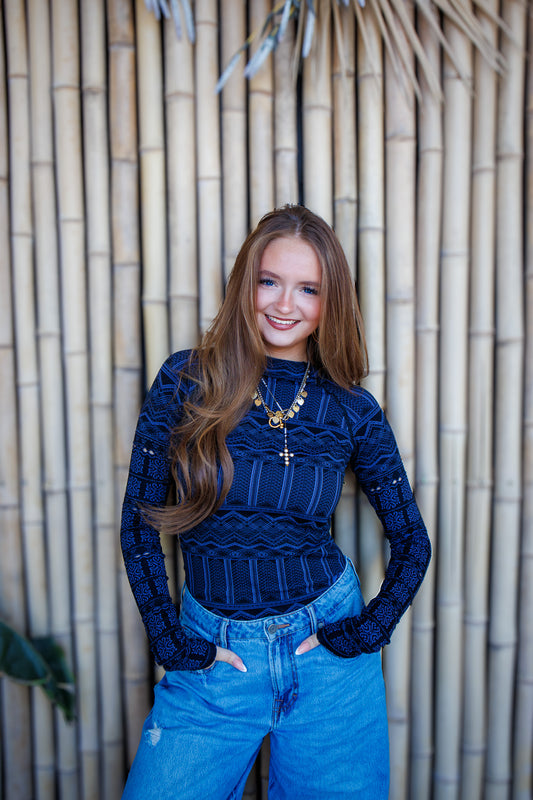 Woman wearing a blue ribbed sweater and jeans standing against a bamboo wall.