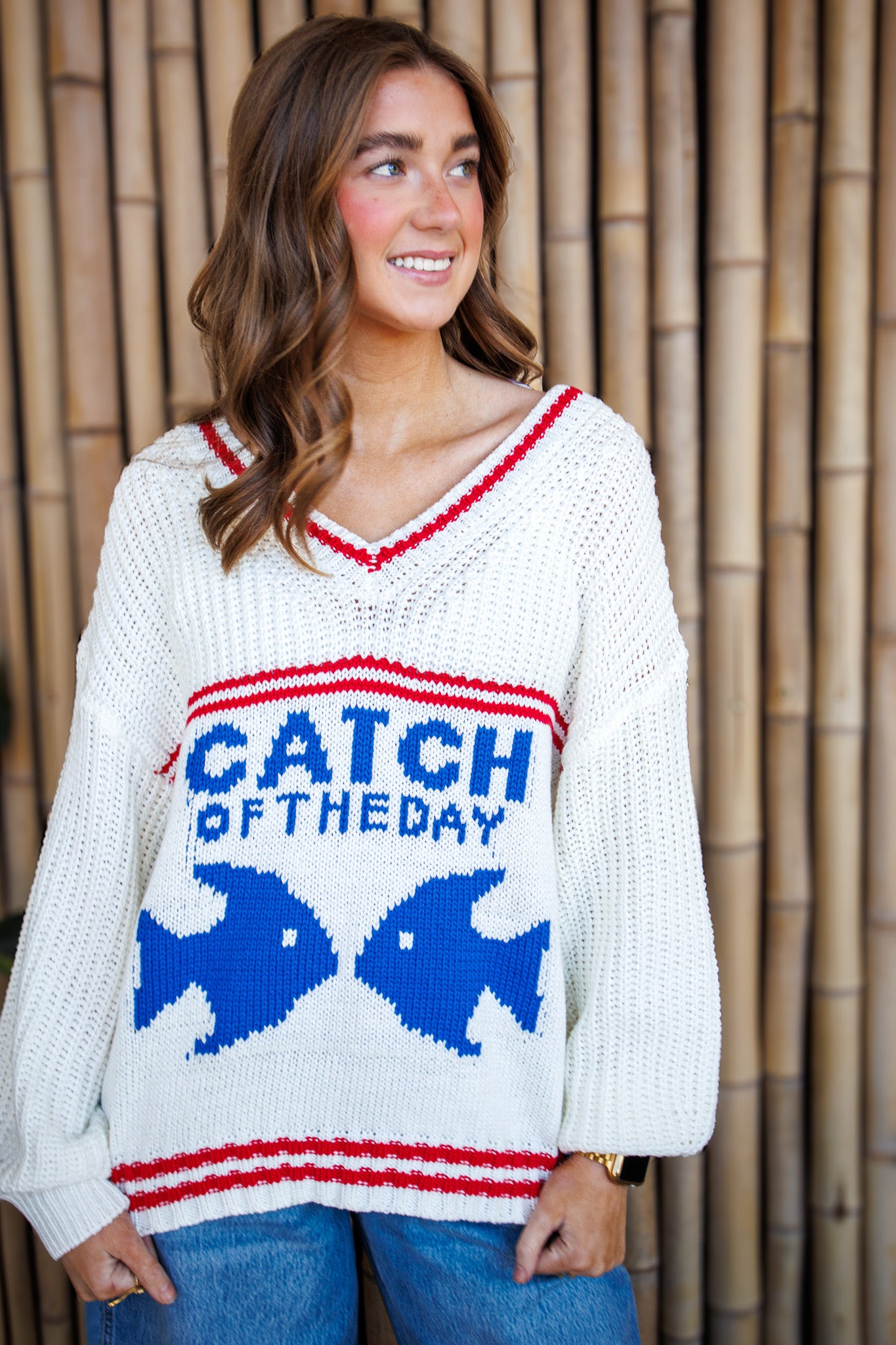 Woman wearing a sweater with fish graphics and text, standing against a wooden paneled background.