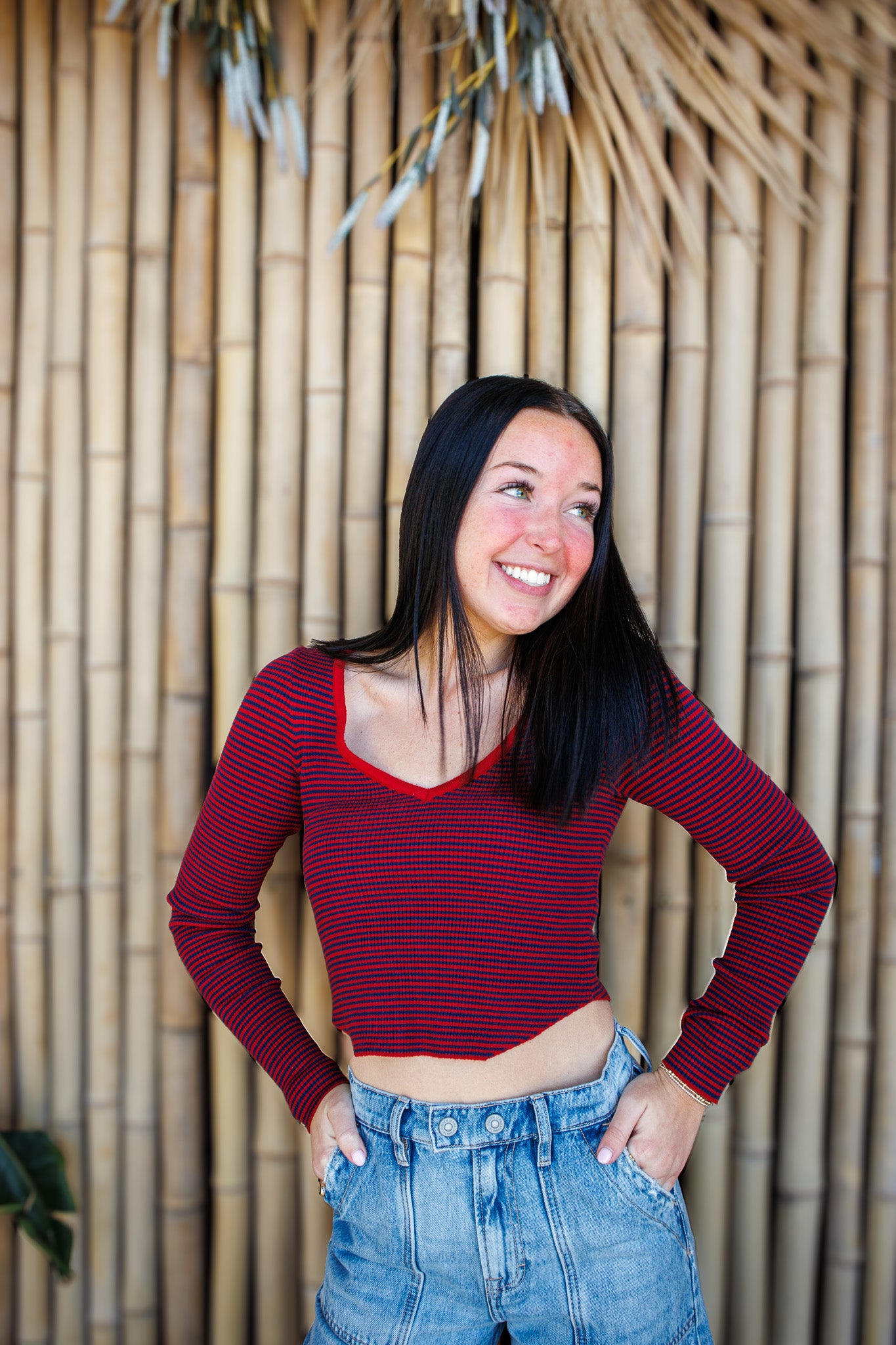 Woman wearing a red striped long-sleeve crop top and blue jeans standing against a bamboo wall.
