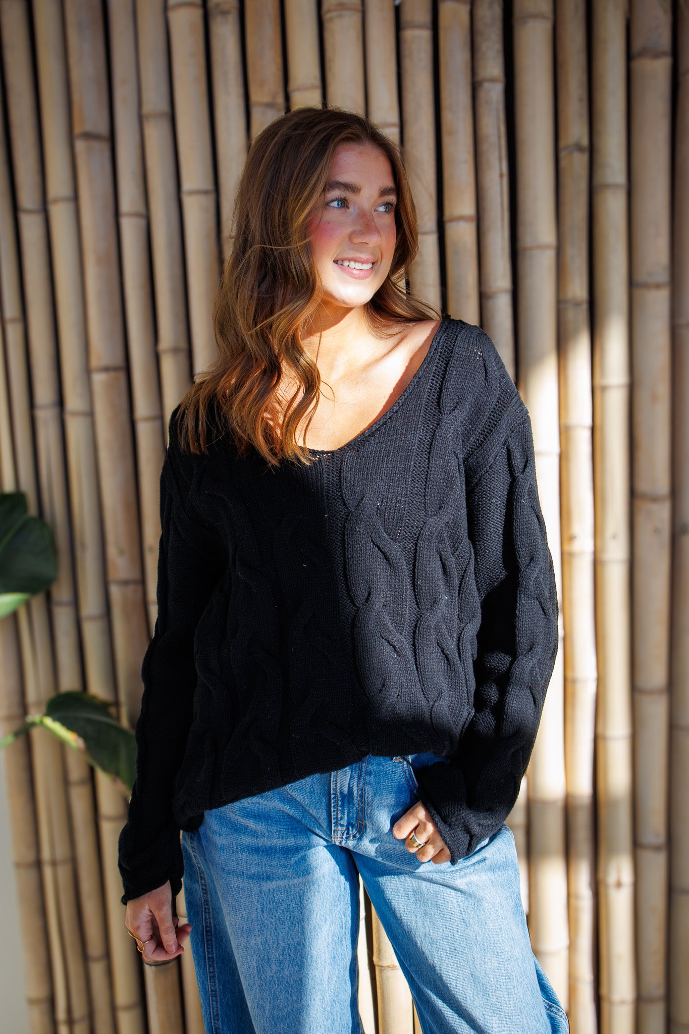 Woman wearing a black sweater and blue jeans standing against a bamboo wall.