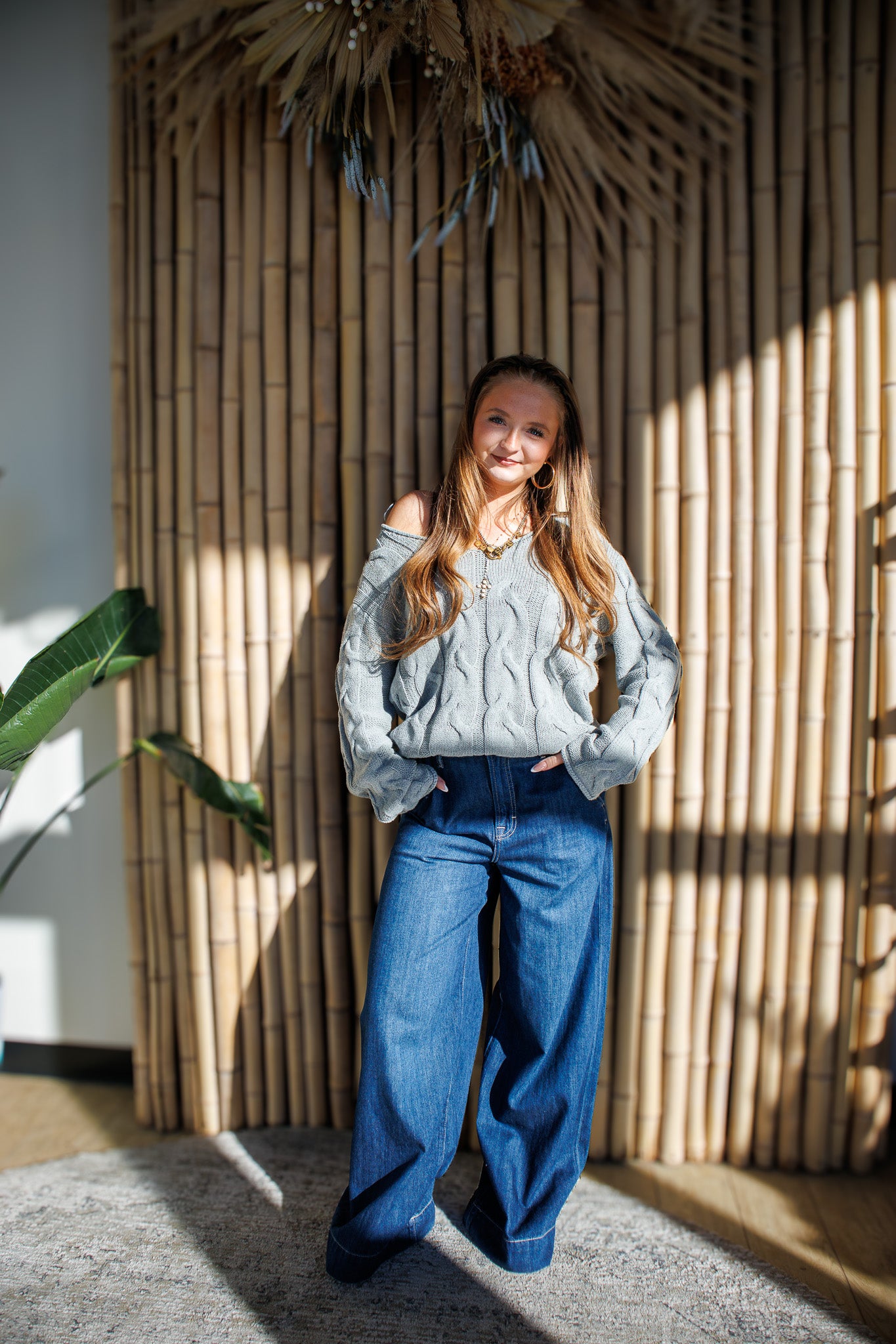 Woman wearing a gray sweater and blue jeans standing in front of a bamboo wall.