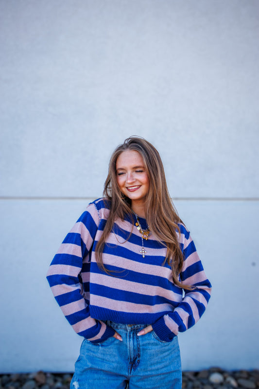 Woman wearing a blue and pink striped sweater and blue jeans against a light gray wall.