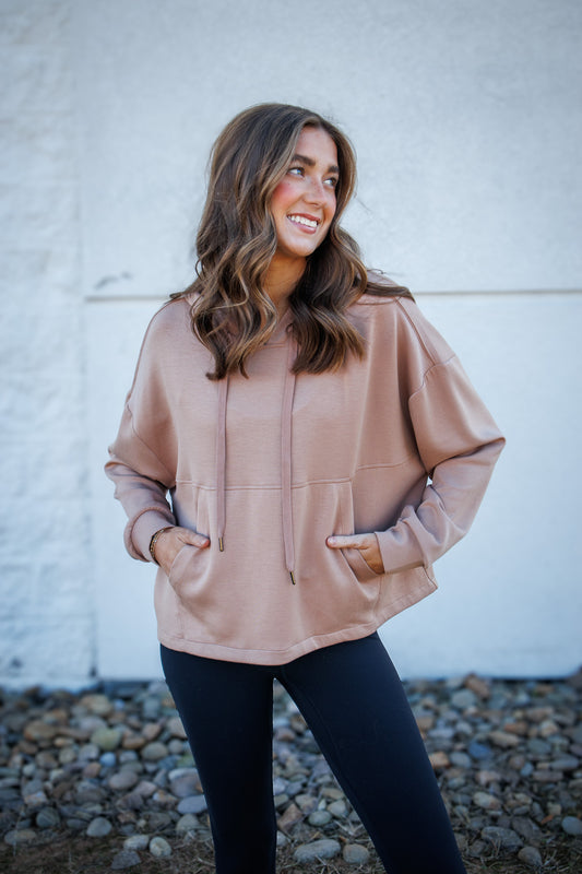 Woman wearing a beige hoodie standing against a white wall with pebbles on the ground.