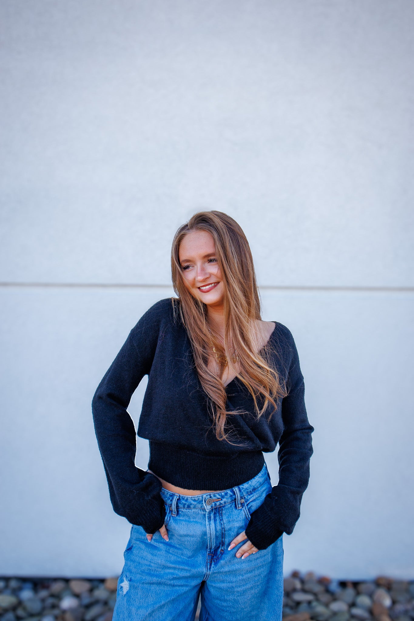 Woman wearing a black sweater and blue jeans standing against a light-colored wall.