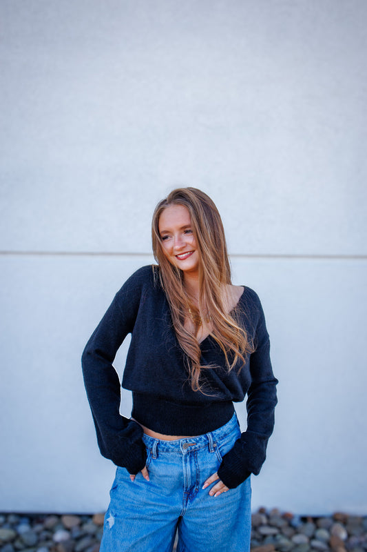 Woman wearing a black sweater and blue jeans standing against a light-colored wall.