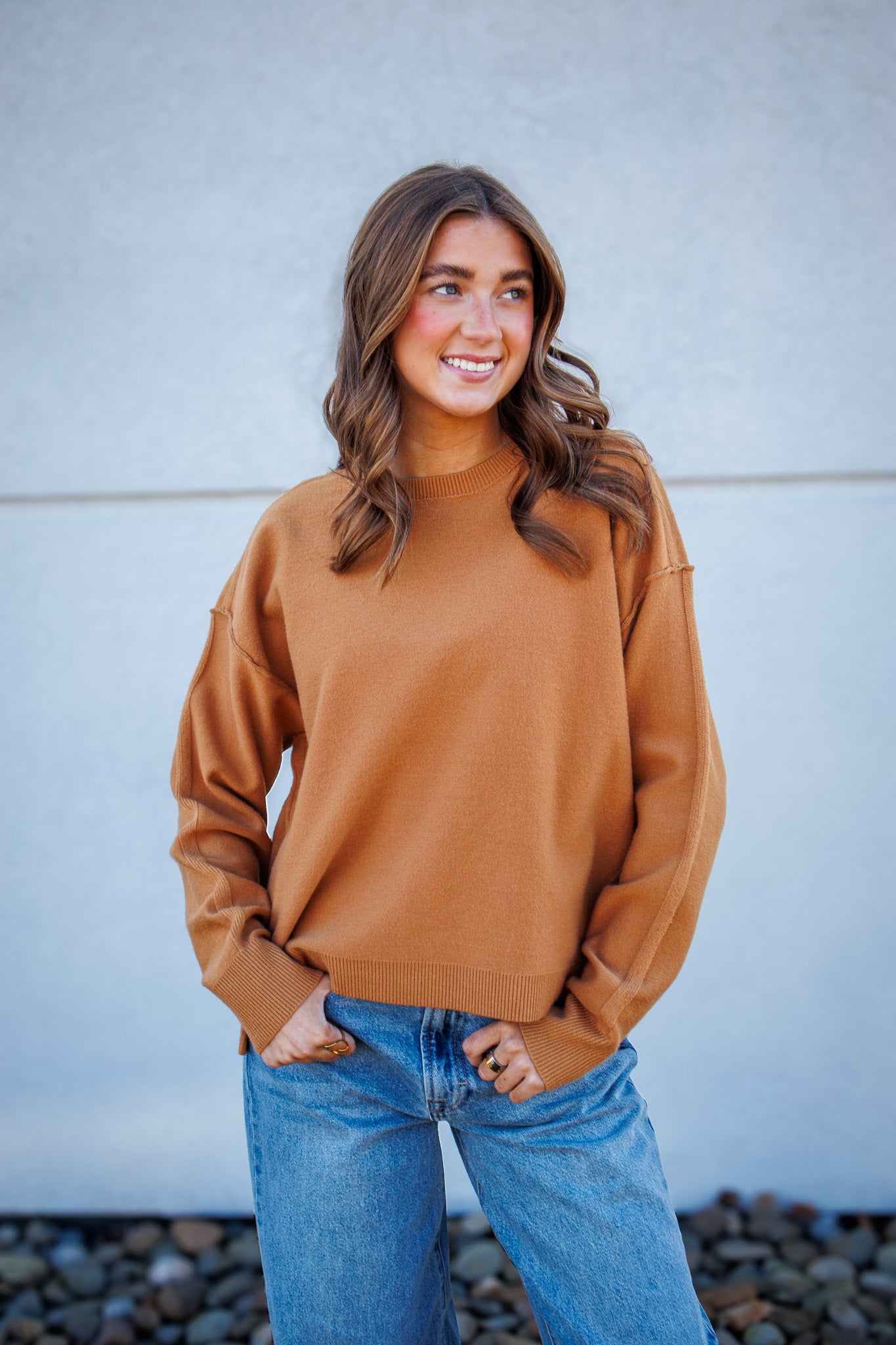 Woman wearing a brown sweater and blue jeans standing against a light gray wall.