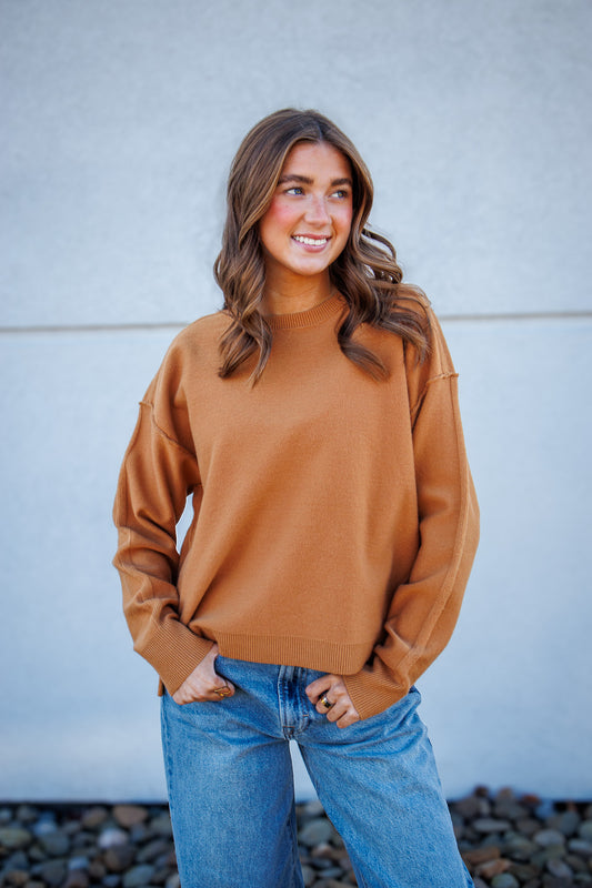 Woman wearing a brown sweater and blue jeans standing against a light gray wall.