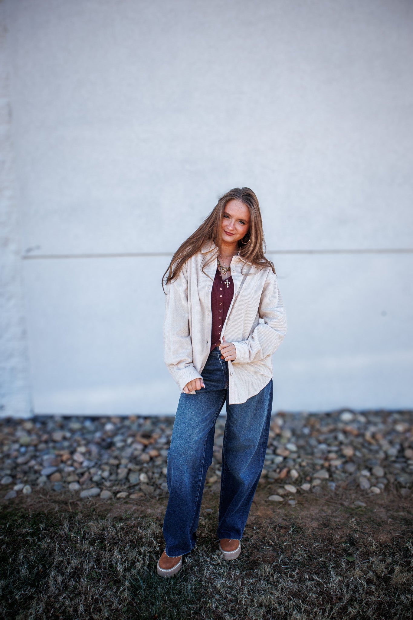 Woman standing outdoors against a plain wall wearing a beige jacket and blue jeans.