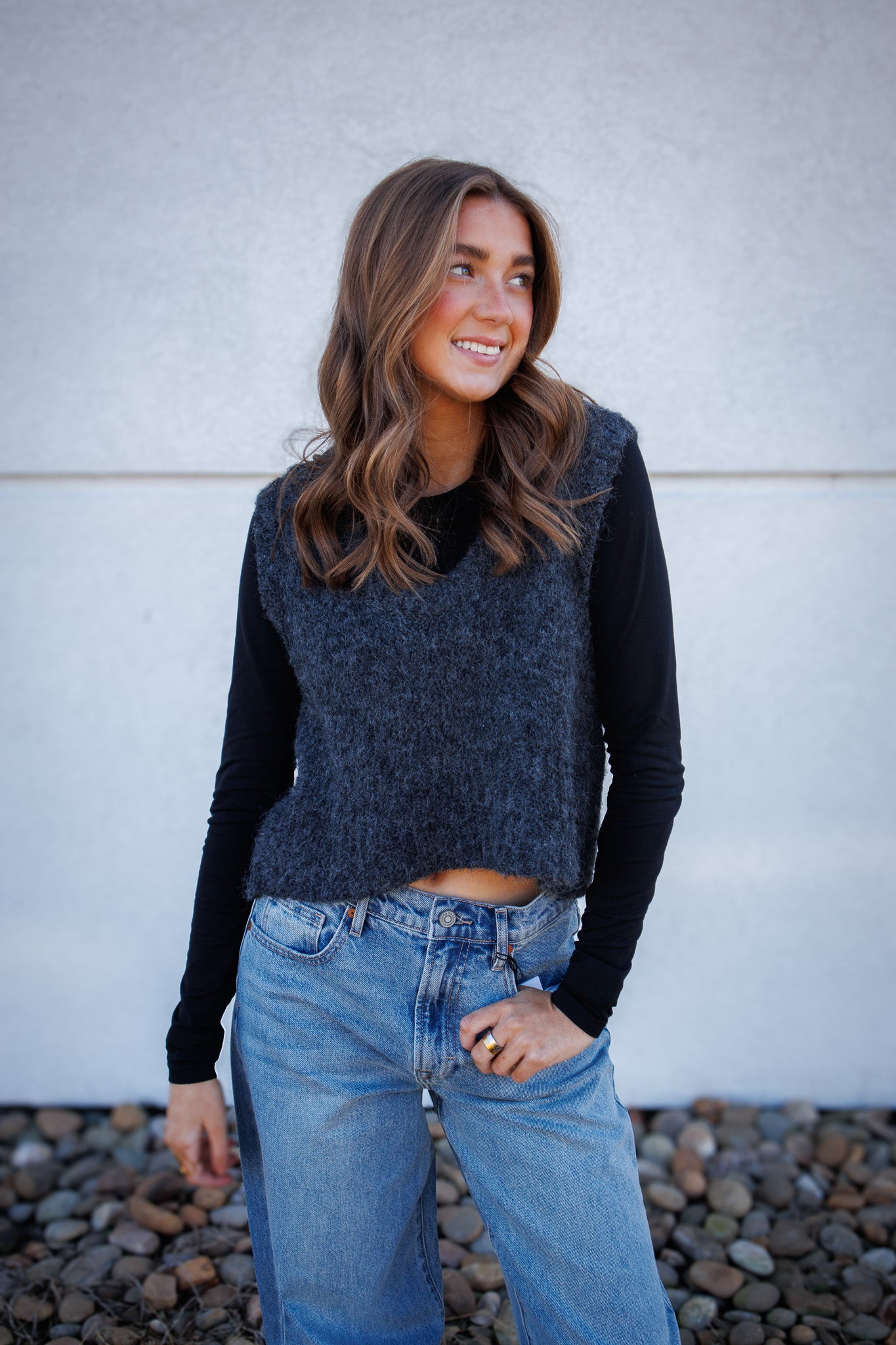 Woman wearing a dark blue sweater and light blue jeans against a gray wall.