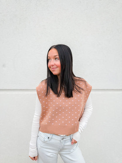 Woman wearing a beige polka dot vest over a white long-sleeve shirt with light wash denim jeans against a white background.