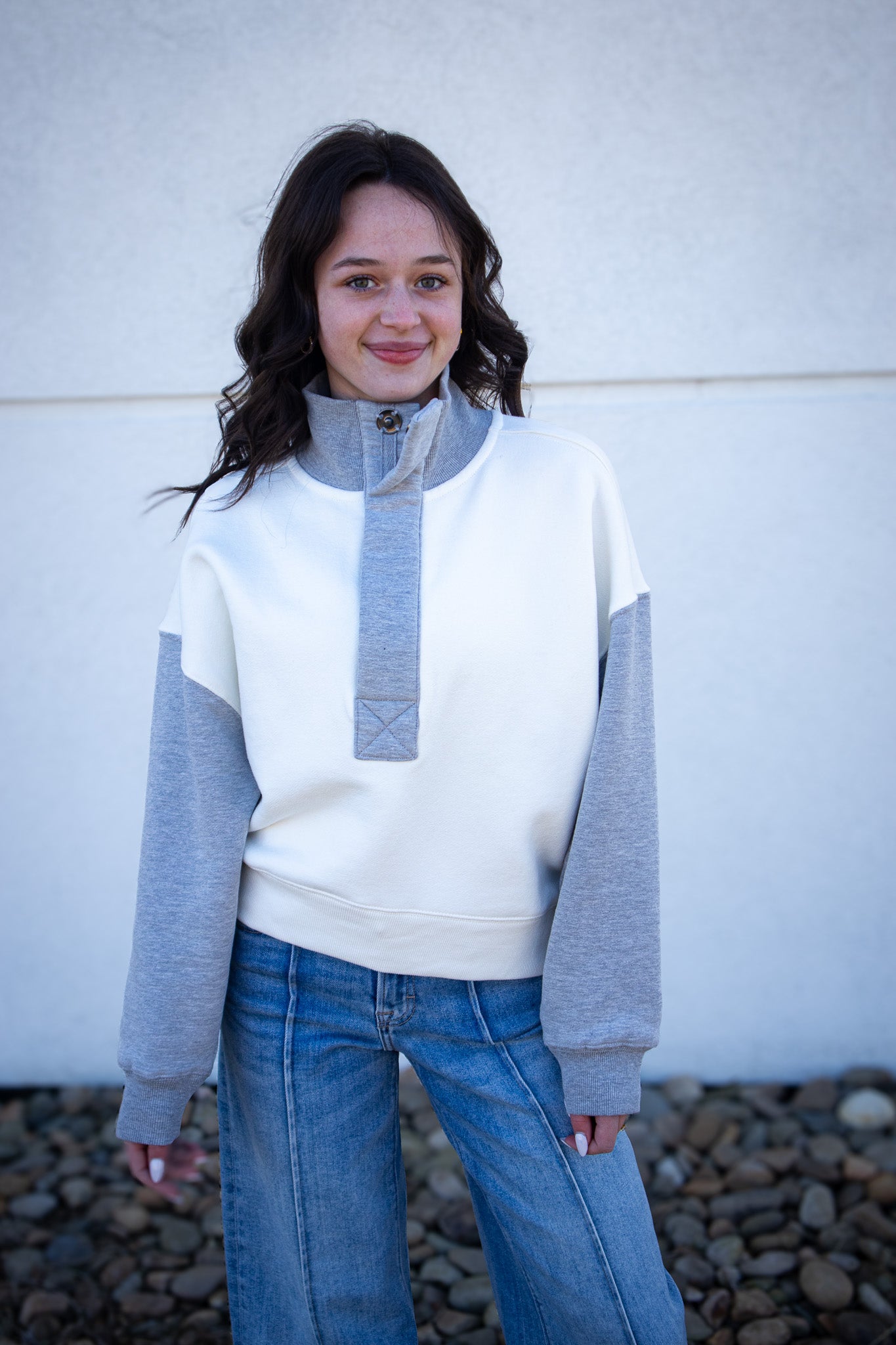 Woman wearing a white and gray sweatshirt with blue jeans against a white wall