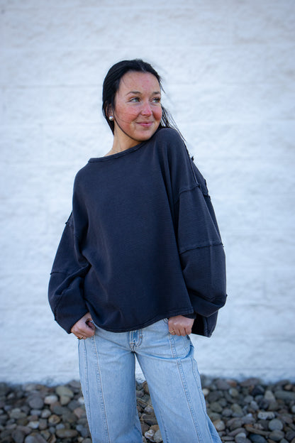 Woman wearing a dark grey oversized sweater and light blue jeans against a white brick wall.
