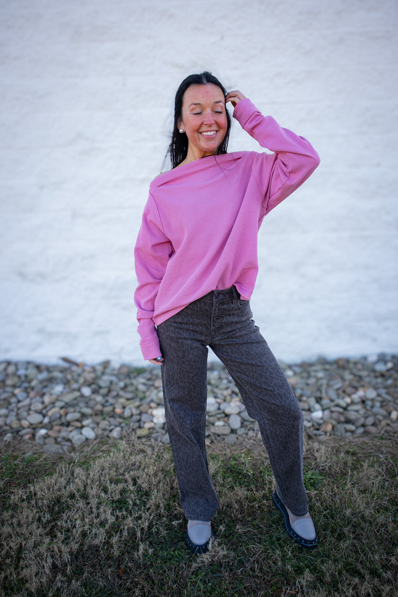 Person wearing a pink sweater and gray pants standing outdoors against a white wall.