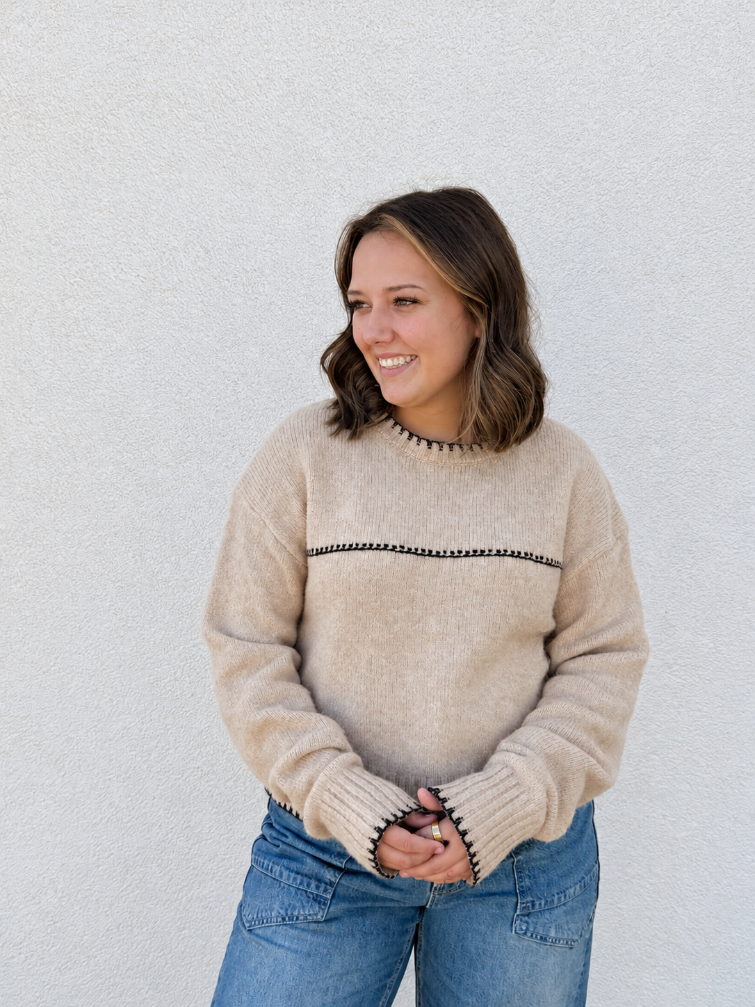 Model wearing the In The Details Contrast Stitch Sweater in Tan & Black, featuring a soft knit fabric, relaxed fit, crew neckline, black contrast stitching, long drop-shoulder sleeves, and ribbed cuffs and hem, styled with light-wash denim jeans.