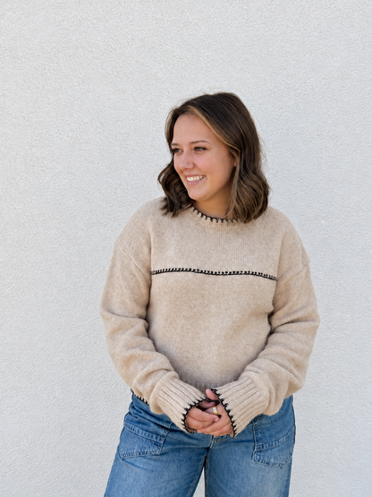 Model wearing the In The Details Contrast Stitch Sweater in Tan & Black, featuring a soft knit fabric, relaxed fit, crew neckline, black contrast stitching, long drop-shoulder sleeves, and ribbed cuffs and hem, styled with light-wash denim jeans.