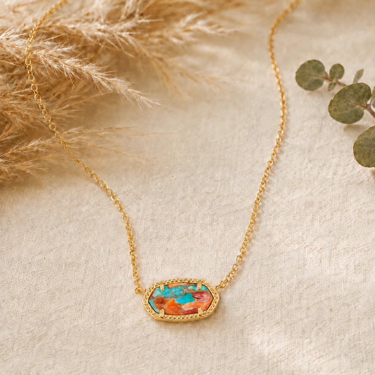 A delicate gold chain necklace with an oval bezel pendant featuring a vibrant turquoise and orange stone inlay, styled on a textured beige fabric with dried pampas grass and eucalyptus leaves.