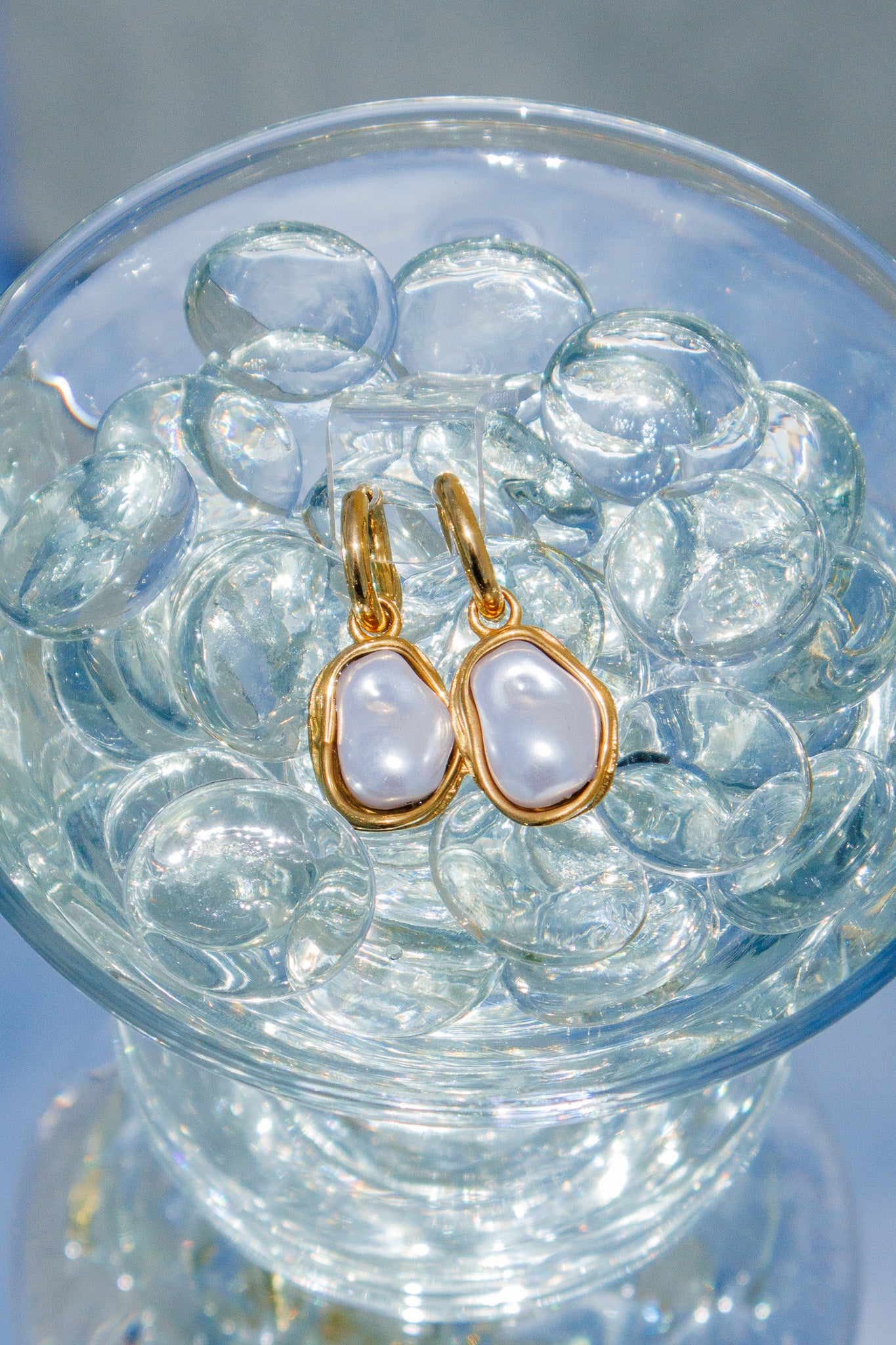 Gold earrings with white stones in a clear glass bowl on a reflective surface
