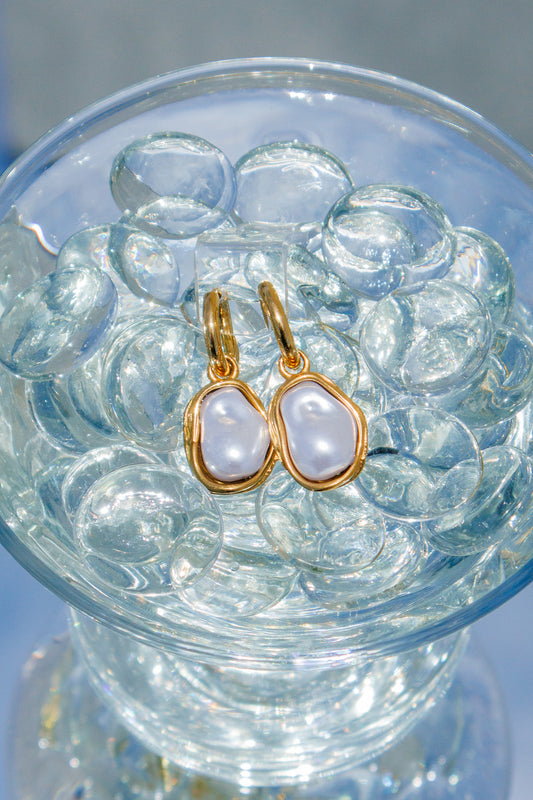 Gold earrings with white stones in a clear glass bowl on a reflective surface