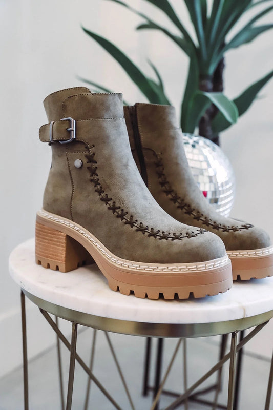 Khaki faux suede ankle boot featuring decorative whipstitch detailing, an adjustable buckle strap, chunky block heel, platform lug sole, and round toe, displayed on a marble table for a stylish fall outfit.