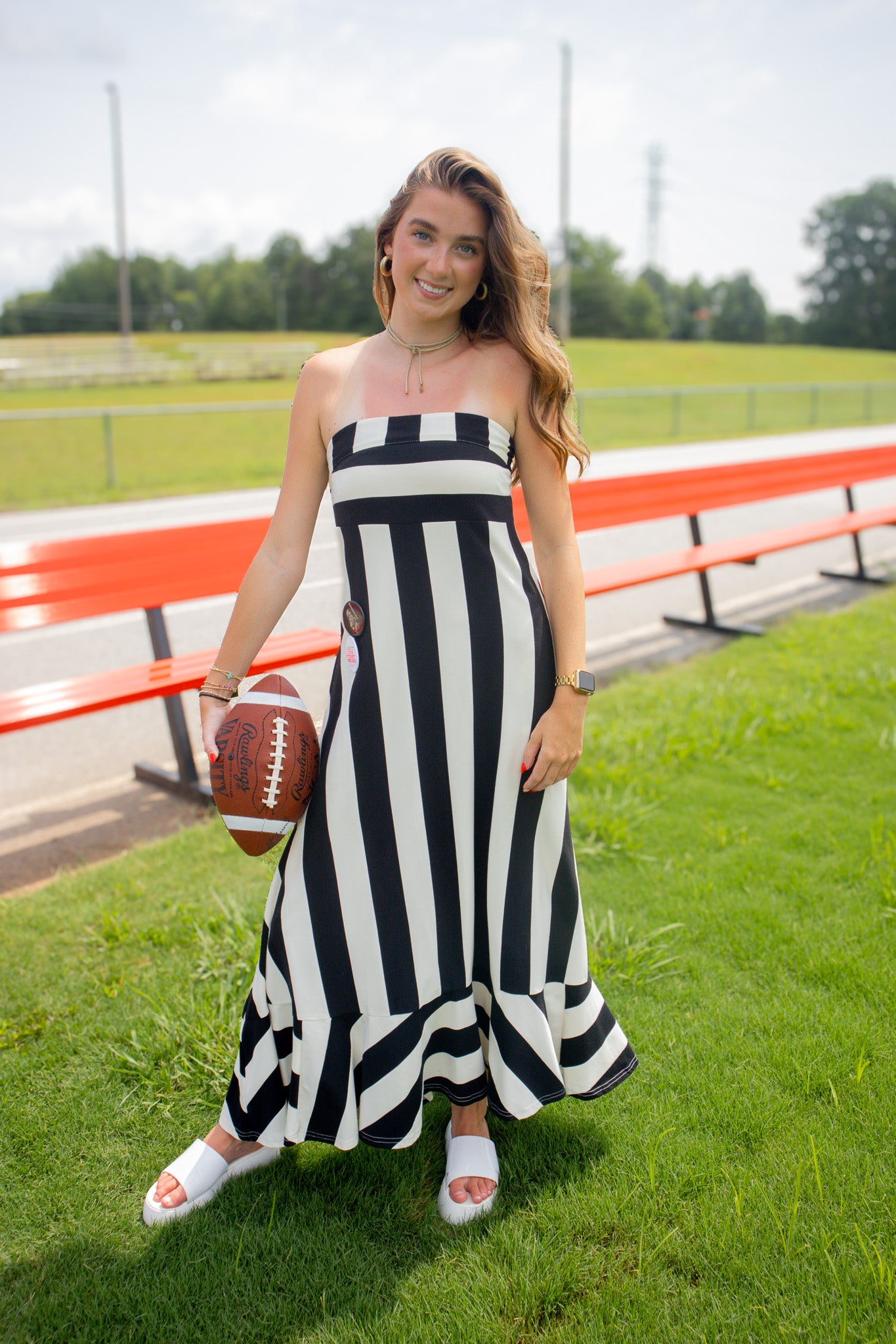 A woman stands on a grassy field near red bleachers, wearing a black and white vertically striped strapless maxi dress and white slide sandals, while holding a brown football.