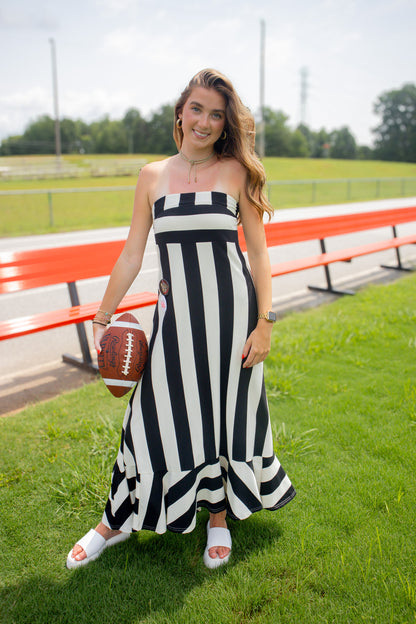 A woman stands on a grassy field near red bleachers, wearing a black and white vertically striped strapless maxi dress and white slide sandals, while holding a brown football.