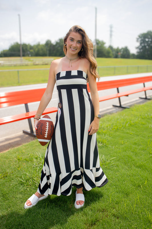 A woman stands on a grassy field near red bleachers, wearing a black and white vertically striped strapless maxi dress and white slide sandals, while holding a brown football.