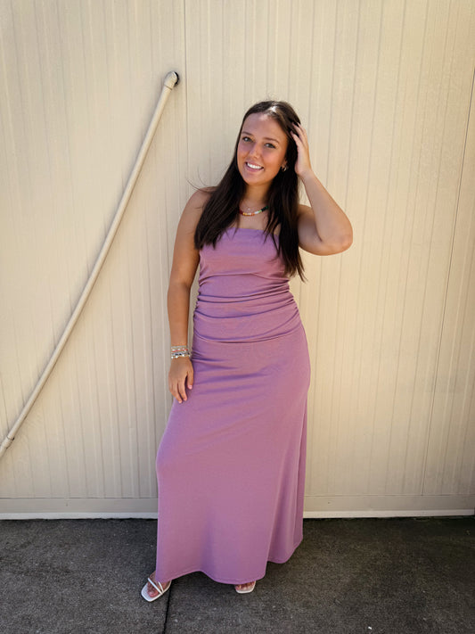 Model wearing a mauve strapless maxi dress featuring a fitted ruched bodice, soft stretchy fabric, and a flowy full-length skirt paired with white sandals for an effortless everyday look.
