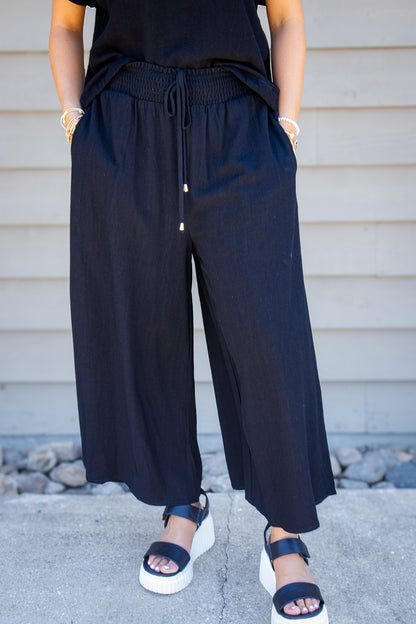 Women’s black linen blend wide leg pants with smocked drawstring waistband and side pockets styled with matching black top and sandals.