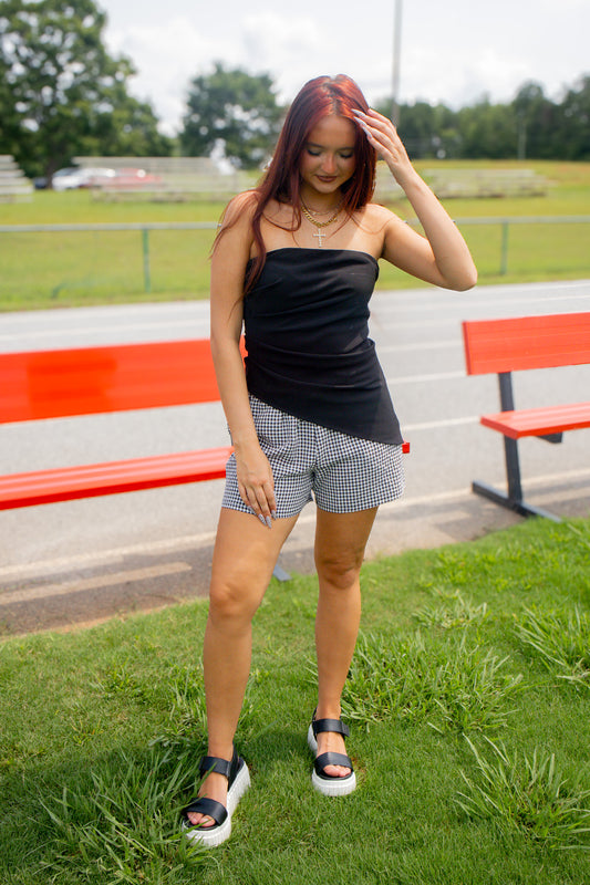 A woman standing on grass outdoors near red benches, wearing a black strapless top, black-and-white gingham shorts, and black platform sandals.