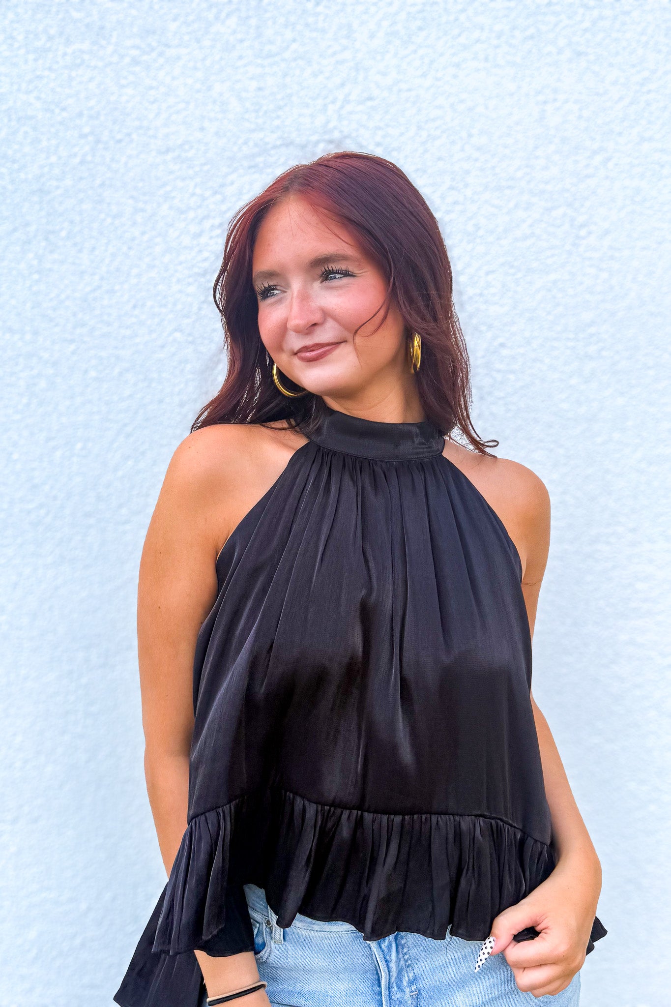 A woman wearing a black halter-neck top with a ruffled hem, paired with light blue jeans, standing against a light blue textured wall.