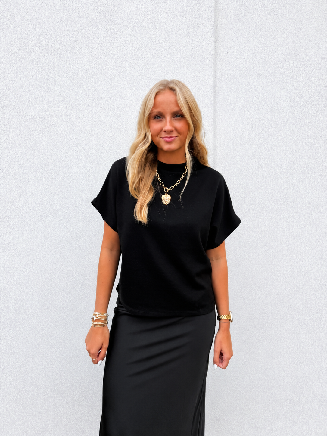 A blonde woman wearing a black short-sleeve mock neck top and a black midi skirt, accessorized with gold necklaces and bracelets, standing against a light gray textured wall.