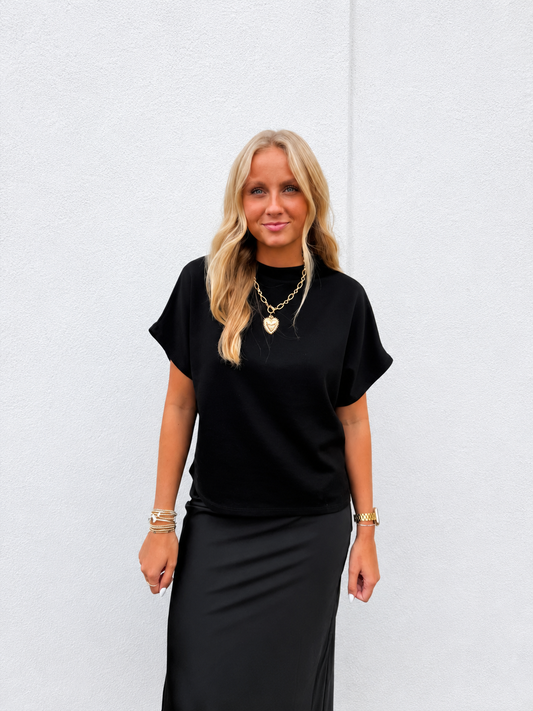 A blonde woman wearing a black short-sleeve mock neck top and a black midi skirt, accessorized with gold necklaces and bracelets, standing against a light gray textured wall.