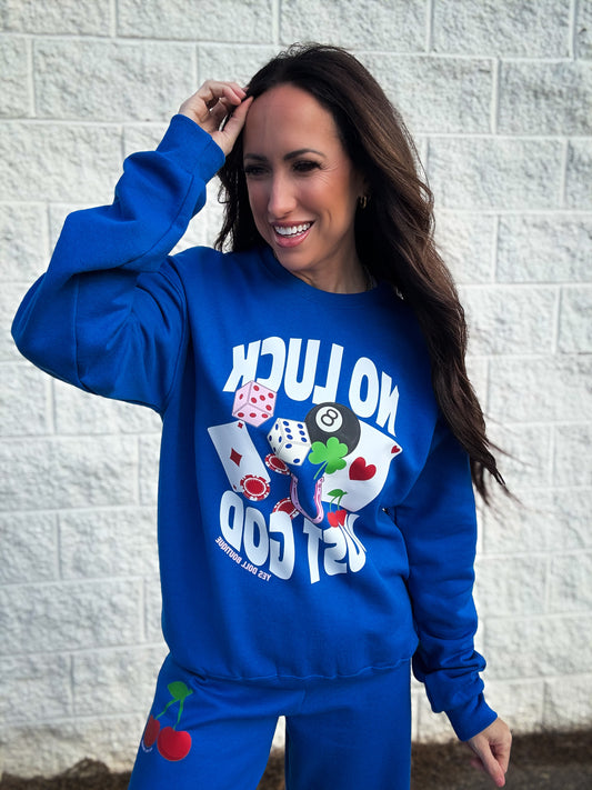 Woman wearing a blue sweatshirt that says 'No Luck Just God' with colorful graphics against a gray brick wall.