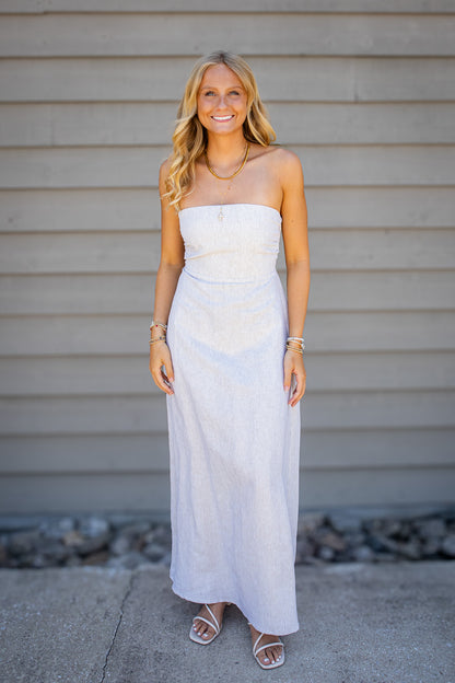 Women’s neutral striped strapless maxi dress styled with white sandals and gold jewelry for a coastal summer outfit.