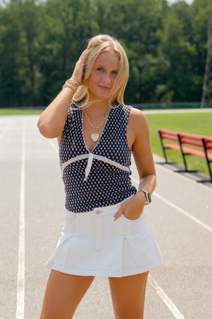 Model wearing the Sideline White Denim Skort featuring a high-rise fit, pleated front, contrast stitching, and built-in shorts, paired with a black and white sleeveless knit top. Perfect for game day. 