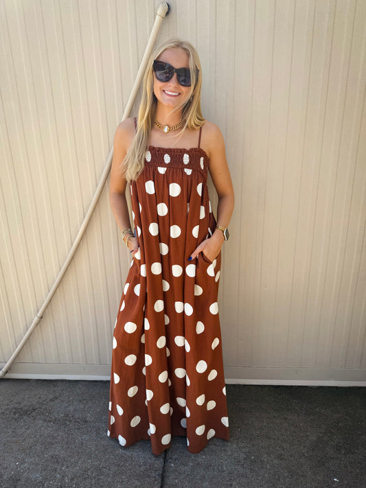 Model wearing a brown maxi dress with oversized ivory polka dots, featuring adjustable spaghetti straps, a smocked neckline, side pockets, and a flowy full-length silhouette paired with sunglasses and gold accessories.